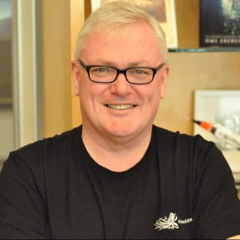 A man wearing black-rimmed glasses and a black T-shirt smiles as he poses for a photo.