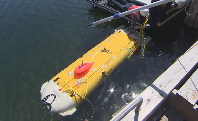 A yellow underwater vehicle floats just at the top of a body of water, as a man wearing a red T-shirt and a white hard hat lays on a platform next to it, tying up the vehicle.