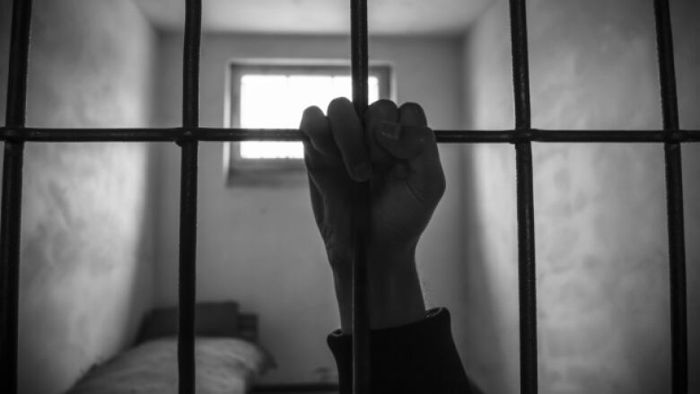 A person's hand is pictured hanging on to metal bars in a prison cell.