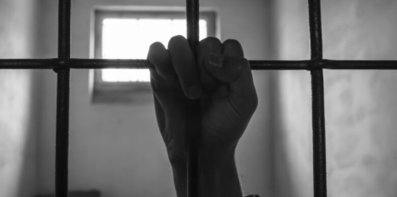 A person's hand is pictured hanging on to metal bars in a prison cell.