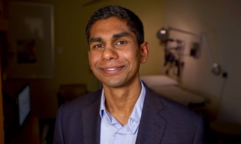 A man with dark hair, wearing a navy blazer and light-blue shirt, sits in a medical office.