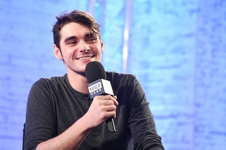 A dark haired man in a grey shirt smiles while holding a microphone.