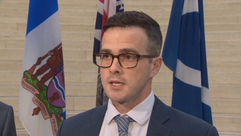 A man wearing a suit and brown-rimmed eyeglasses stands in front of three flags.