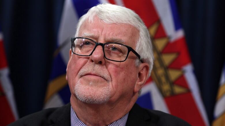 A man with black glasses and white hair looks into the distance. Behind him is a flag of British Columbia.