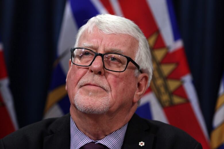 A man with black glasses and white hair looks into the distance. Behind him is a flag of British Columbia.