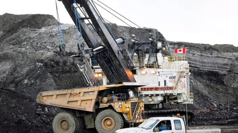 A huge white machine with a shovel looks ready to put a load into the back of very large, orange truck. A man in a white pickup truck watches.  