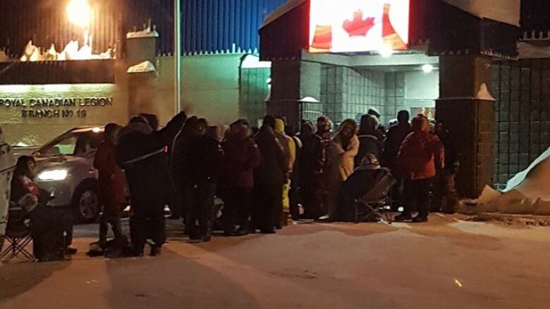 A lineup of people stand outside a building with a Canadian flag hanging from the front. 