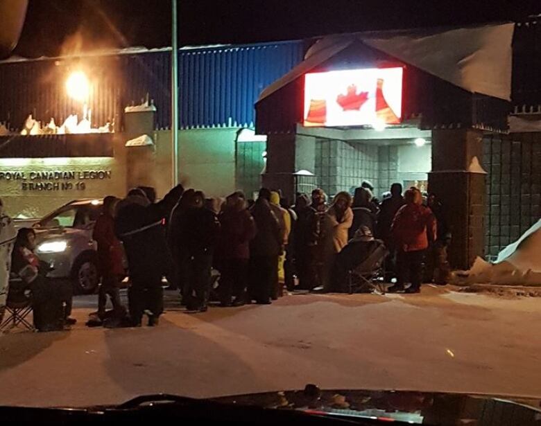 A lineup of people stand outside a building with a Canadian flag hanging from the front. 