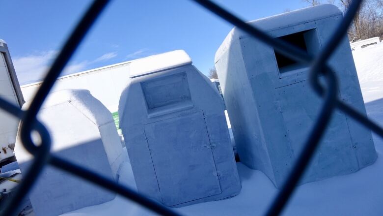 Three cardboard bins painted white are seen through a chainlink fence on a snowy day