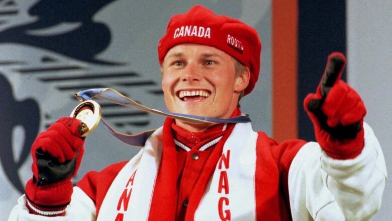 A smiling Olympic athlete dressed in Canadian red and white is shown wearing a gold medal around his neck, and holding the medal up in his right hand.