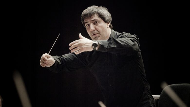 A man with dark hair is shown with his hands in front of him, while conducting an orchestra. The background is dark.