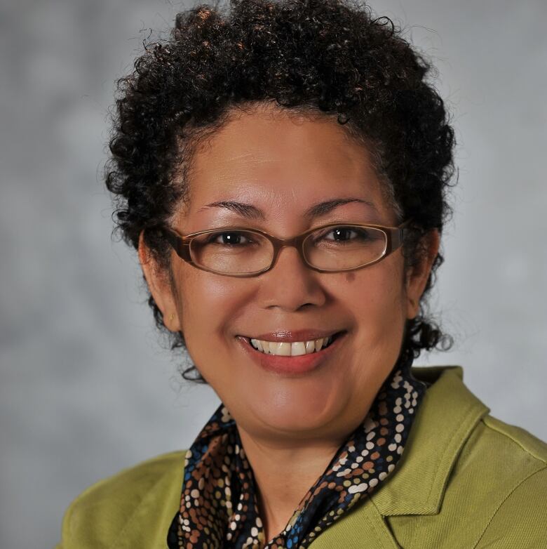 A smiling woman with black curly hair wearing glasses, a green jacket and a thin scarf