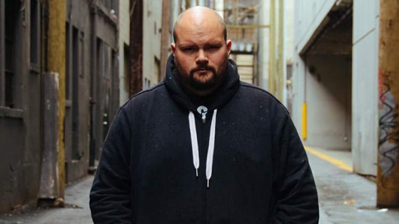 A man wearing a black hoodie with white strings stands in a Vancouver alleyway.