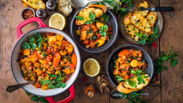 Overhead shot of a red dutch oven with a root vegetable stew on a wooden surface. 2 bowls of the stew and a plate with sliced bread sit next to the dutch oven. 