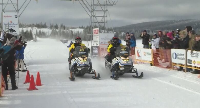 Two snowmobiles ride next to each other through a finish line with fans lining both sides. 