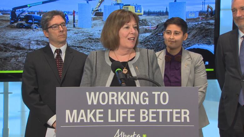  a woman speaks at a microphone behind a sign saying 