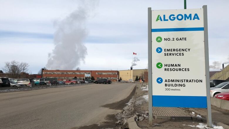 An Algoma steel sign welcomes visitors to the work site while a grey tuft of smoke rises over a brick office building.