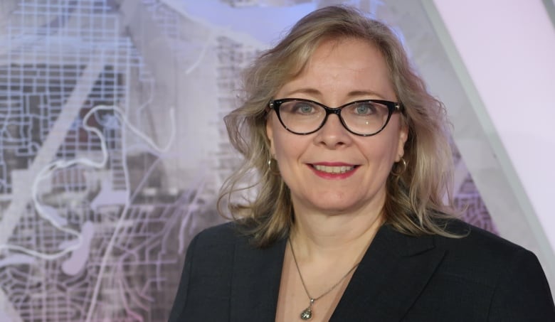 A woman with blonde hair and glasses is smiling in front of a TV set, which shows a map of Calgary. 