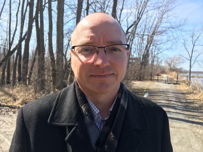 A bald man wearing glasses, a blue and white shirt and a black jacket standing in a forest