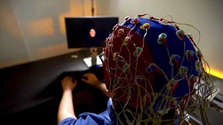 A person wears a cap on their head with sensor nodes and wires while watching a display monitor.