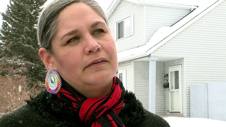 A woman stands in front of her home on a snowy day. She is wearing a red scarf and colourful earrings. 