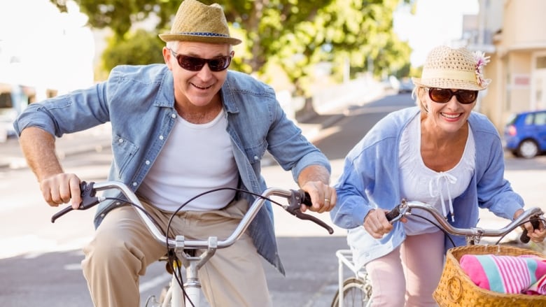 Two seniors wearing sunglasses and hats smile as they ride their bicycles.