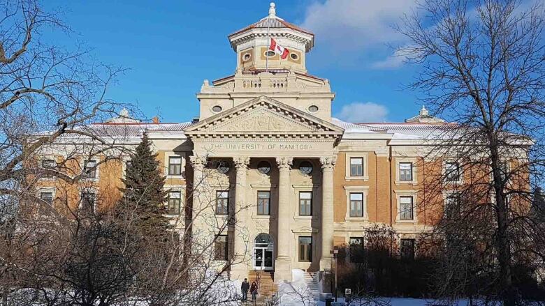 Large neoclassical style orange building with white panneling. A canadian flag hangs from the builing and the building is shrouded by trees. It's winter, and there is snow on the ground and the trees don't have leaves.