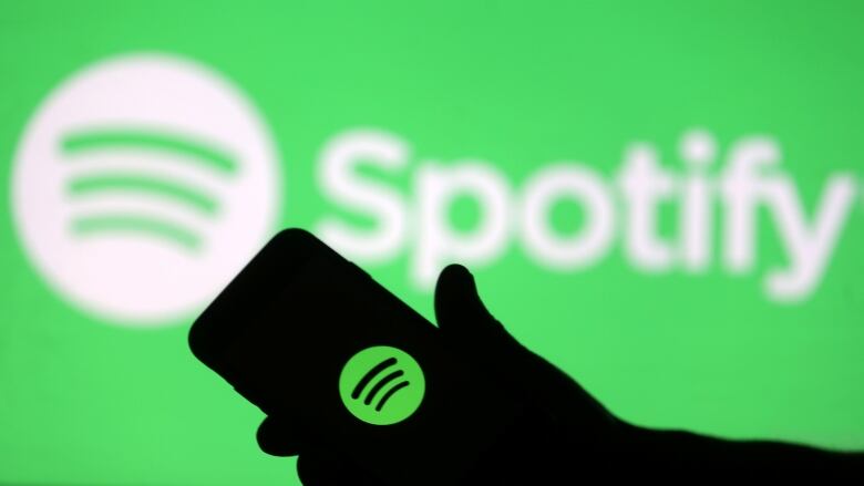 A silhouette of someone's hand holds a phone that displays the Spotify logo. The background is green and also shows the Spotify logo.