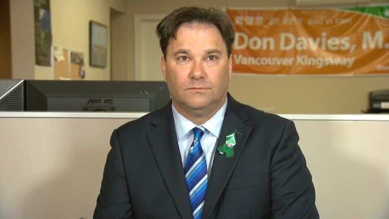 A man in a suit stands in an office. An orange sign hangs in the backgroud. It reads 