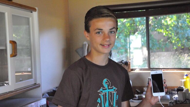 A young boy, his hair neatly combed to the side and wearing a brown t-shirt, holds up his iPhone while grinning at the camera, light from the window behind him filtering into the room.