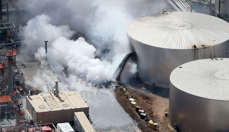 Thick white smoke billows as a black liquid pours out of a large refinery tank.