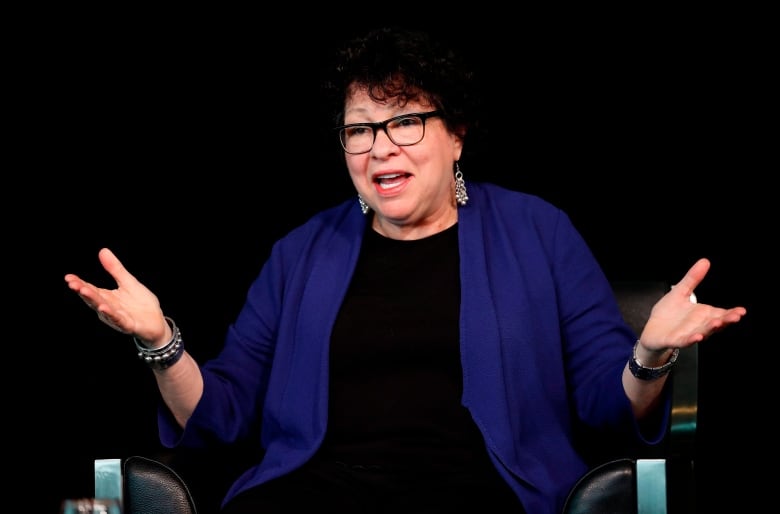 A woman with glasses and curly dark hair smiles and holds up hands