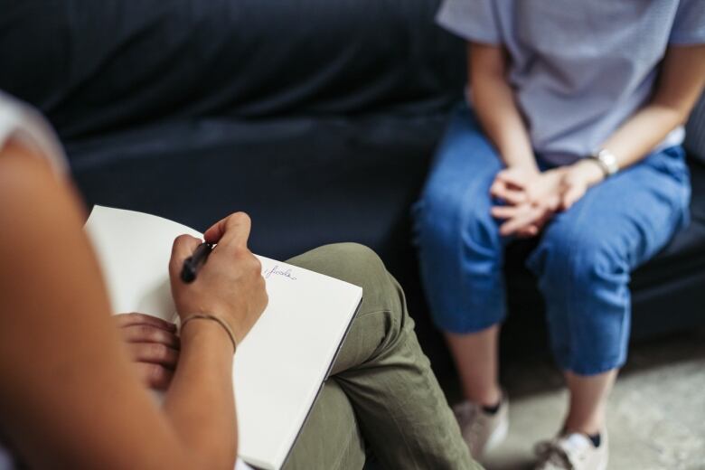 A person sits on a couch facing a person writing in a notebook.