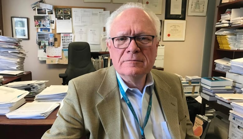 A doctor looks directly into the camera while sitting in his office. There are files stacked on the desk behind him.