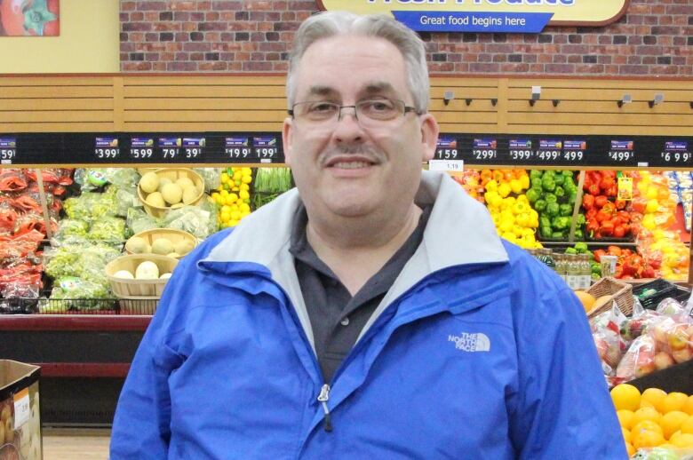 A middle aged man wears a bright jacket inside a grocery store, posing in front of the produce.