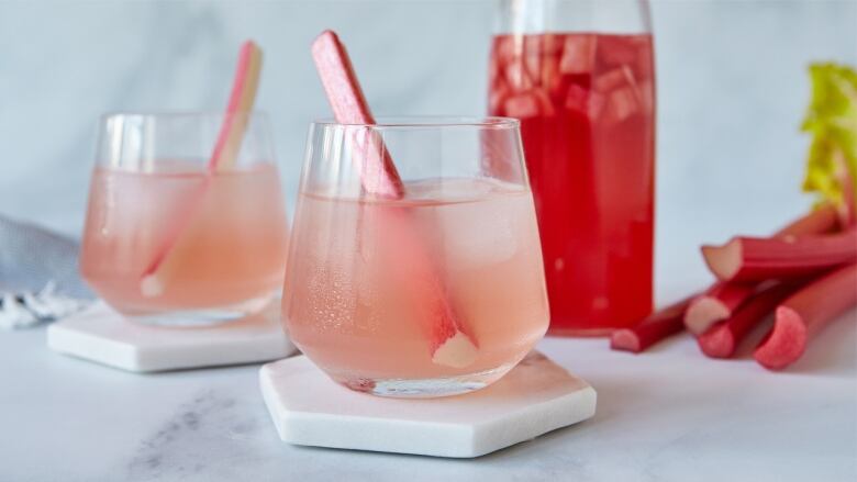 2 glasses on marble coasters on a marble countertop. they have a pink drink in them and a rhubarb stalk in each. a pitcher of bright pink drink sits behind them, with cut rhubarb stalking laying next to it. 