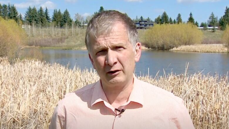 A man in a light shirt is seen standing in daylight with a body of water and shrubs in the background.