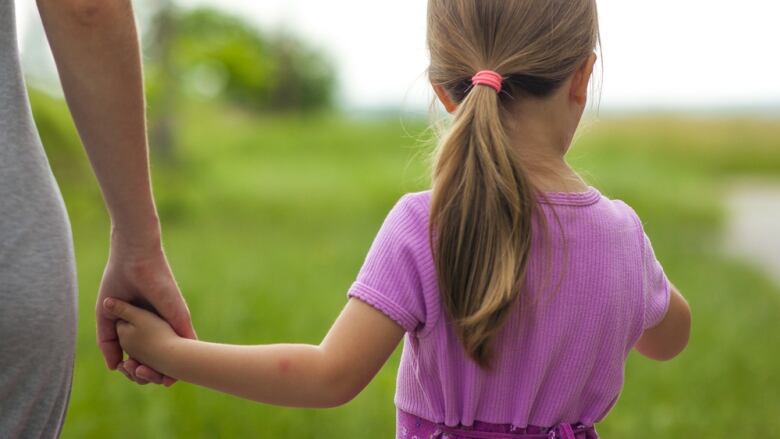 An adult's hand holds the hand of a young girl in a pink shirt, seen from behind.