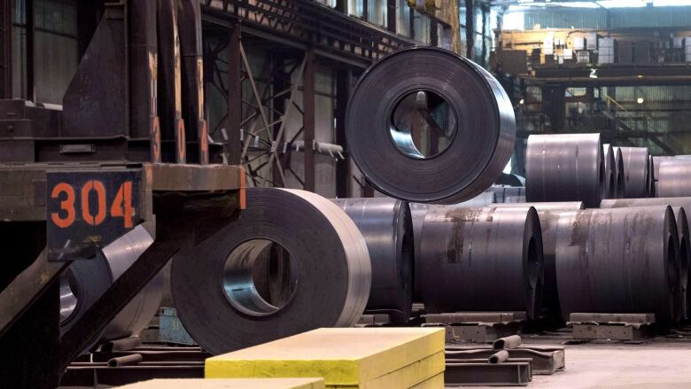 Large steel drums are seen on a factory floor, labelled with numbers and being lifted by a crane.