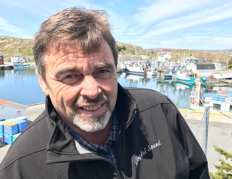 A smiling man with a salt-and-pepper goatee stands in front of a small harbour lined with fishing boats.