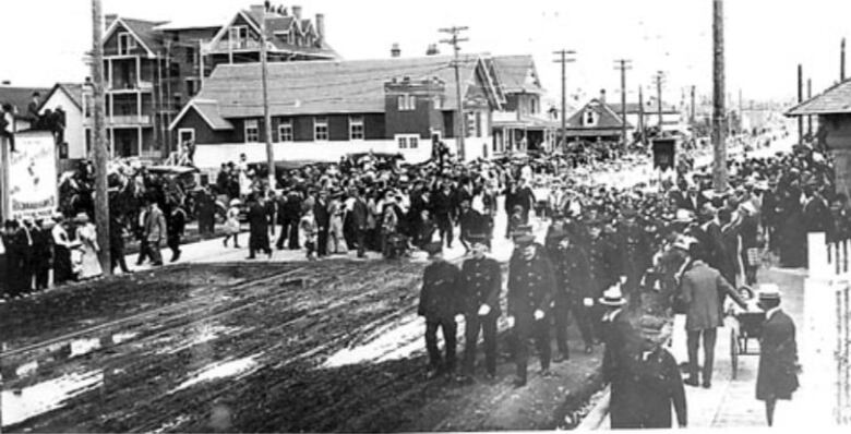 Black and white photo showing a parade of people walking down a dirt street through what appears to be a residential area.