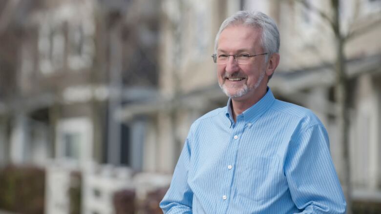 A white man with grey hair and a blue dress shirt stands outside.