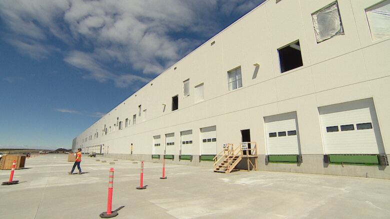 a large warehouse against a blue sky. a worker in a high visibility safety vest walks holding a box.