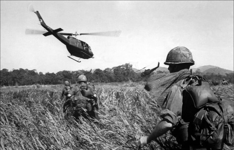 Soldiers walking through high grass while a helicopter flies overhead.