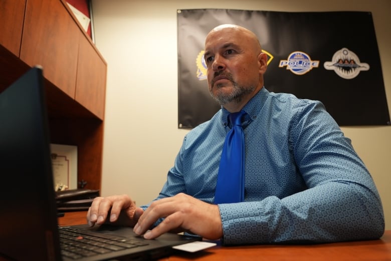 A man in a suit and tie stands types at a computer with police logos visible on a poster behind him.