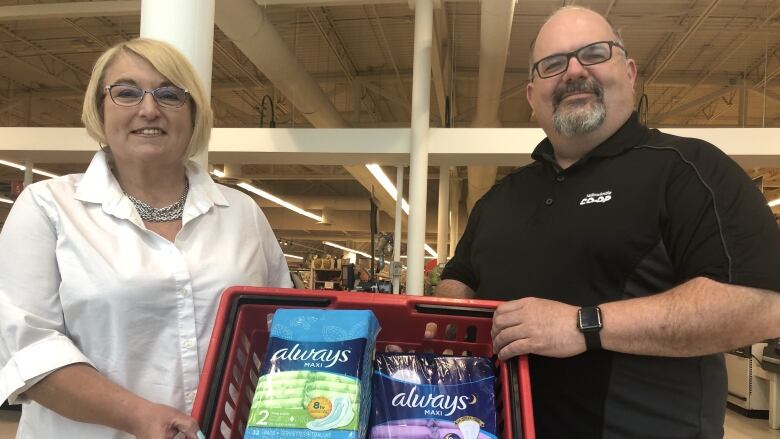 Two people stand holding a bin of tampons. 