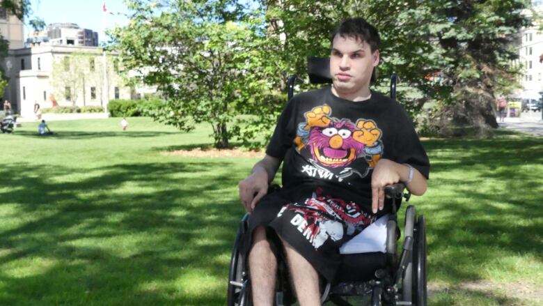 A young man in a wheelchair and wearing a black t-shirt with Animal from the Muppets sits outside in a park on a bright day.