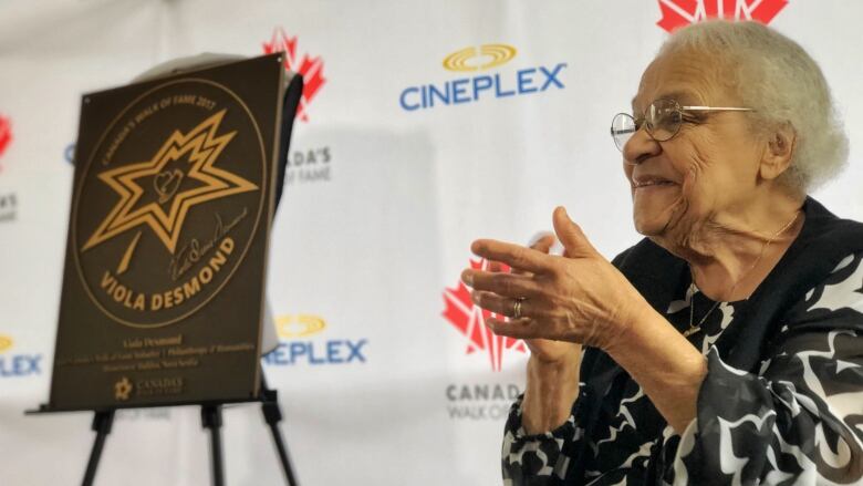 An older Black woman wearing a black and white blouse sits beside a sign that reads 