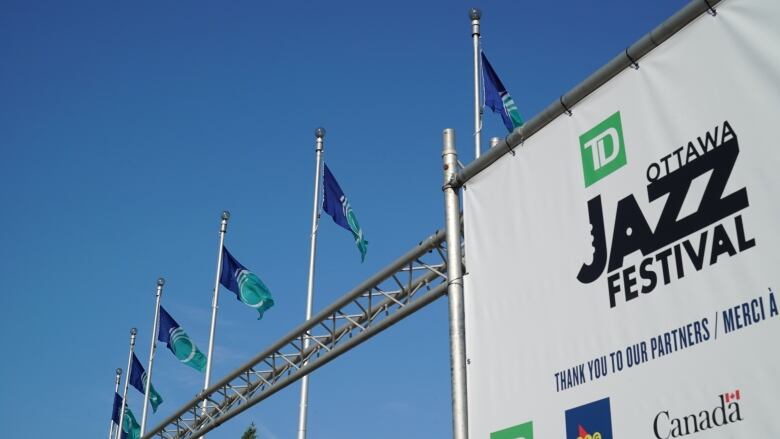 A sign against a bright blue sky. Several City of Ottawa flags fly in the background. The sign reads 