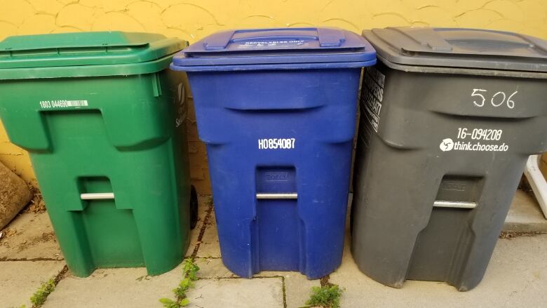 Three garbage bins of the same size stand next to each other on outdoor tile, in front of a yellow wall. The left bin is green, the middle one is blue, and the right bin is grey.